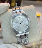 A silver-tone stainless steel watch with a textured white dial and matching metal bracelet, displayed on a beige cushion held by a white-gloved hand in a retail store.