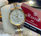 A two-tone SeaStar watch with a gold and silver metal bracelet, gold fluted bezel, and a white dial, held by a white gloved hand. A red SeaStar brand tag is attached to the watch, and a blurred store interior is visible in the background.