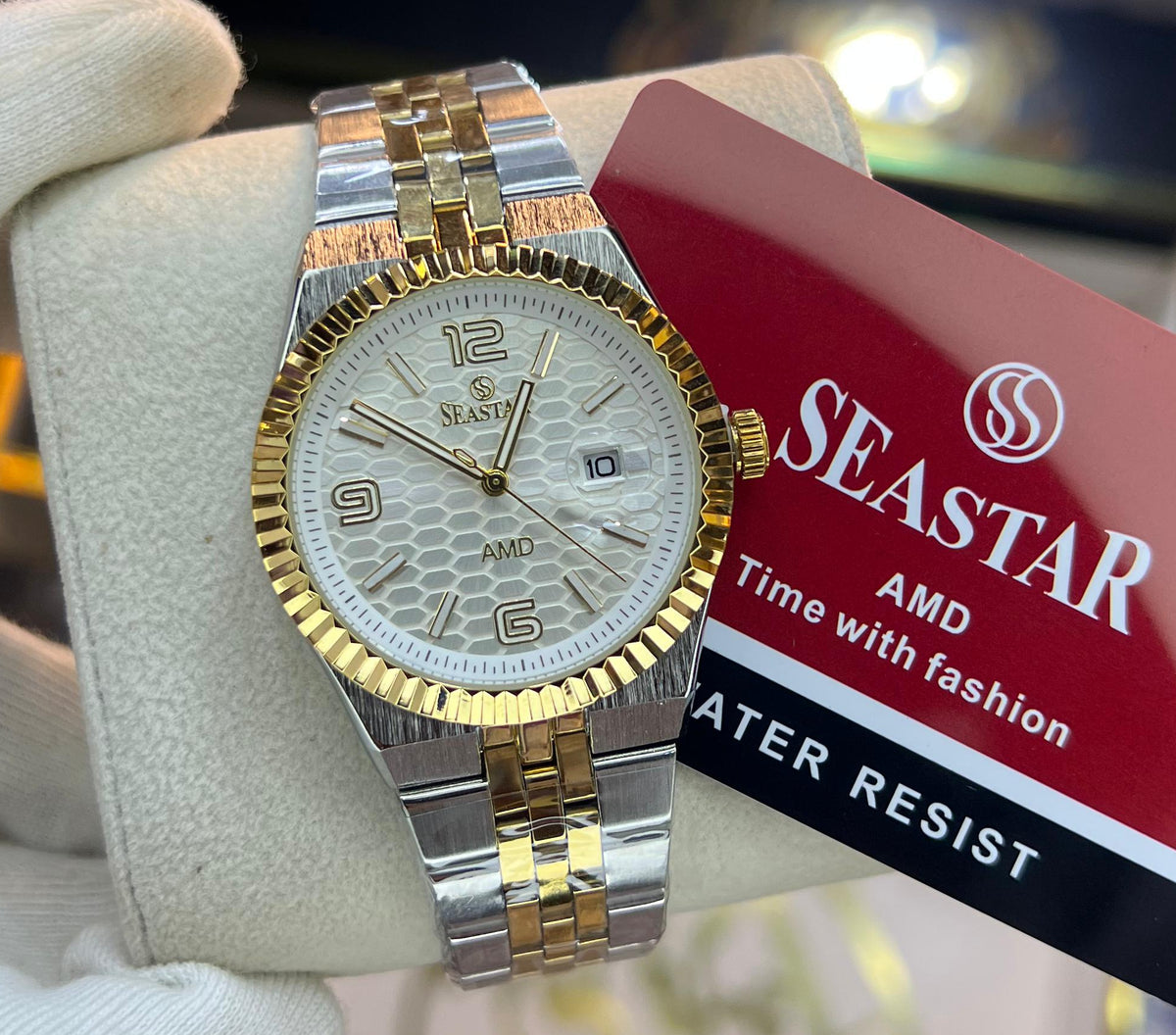 A two-tone SeaStar watch with a gold and silver metal bracelet, gold fluted bezel, and a white dial, held by a white gloved hand. A red SeaStar brand tag is attached to the watch, and a blurred store interior is visible in the background.