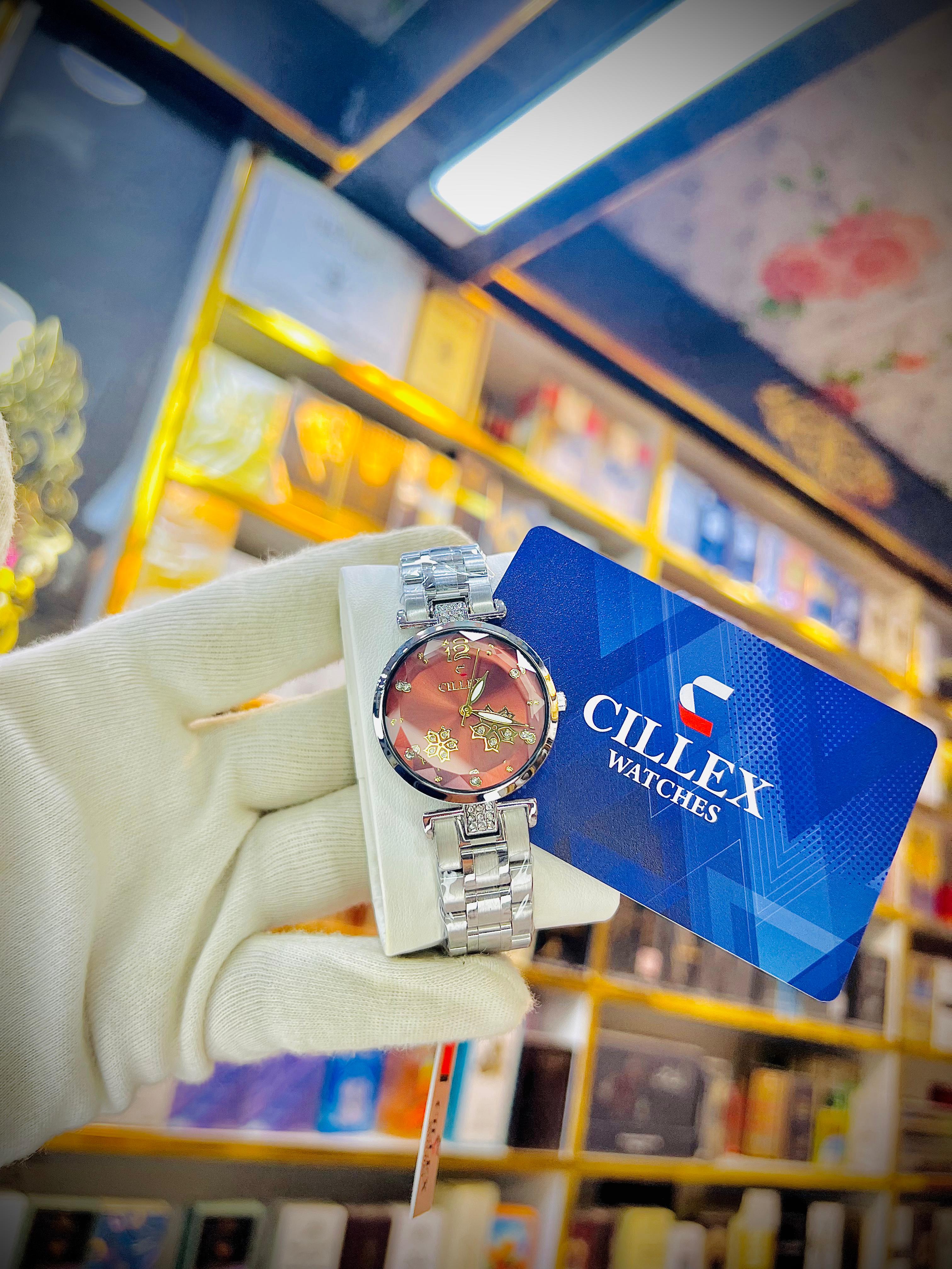 A silver metal link watch with a round rose-gold dial is held in a white-gloved hand, accompanied by a blue brand tag that reads 'CILLEX WATCHES'. The background shows a blurred retail store shelf.