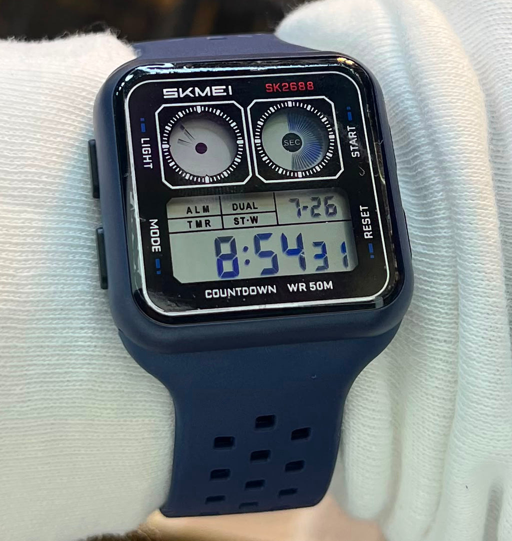 A square digital watch with a dark navy blue strap and black case, worn on a person's wrist covered in a white long-sleeve cuff, with a blurred retail store shelf background.