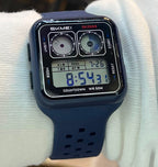 A square digital watch with a dark navy blue strap and black case, worn on a person's wrist covered in a white long-sleeve cuff, with a blurred retail store shelf background.