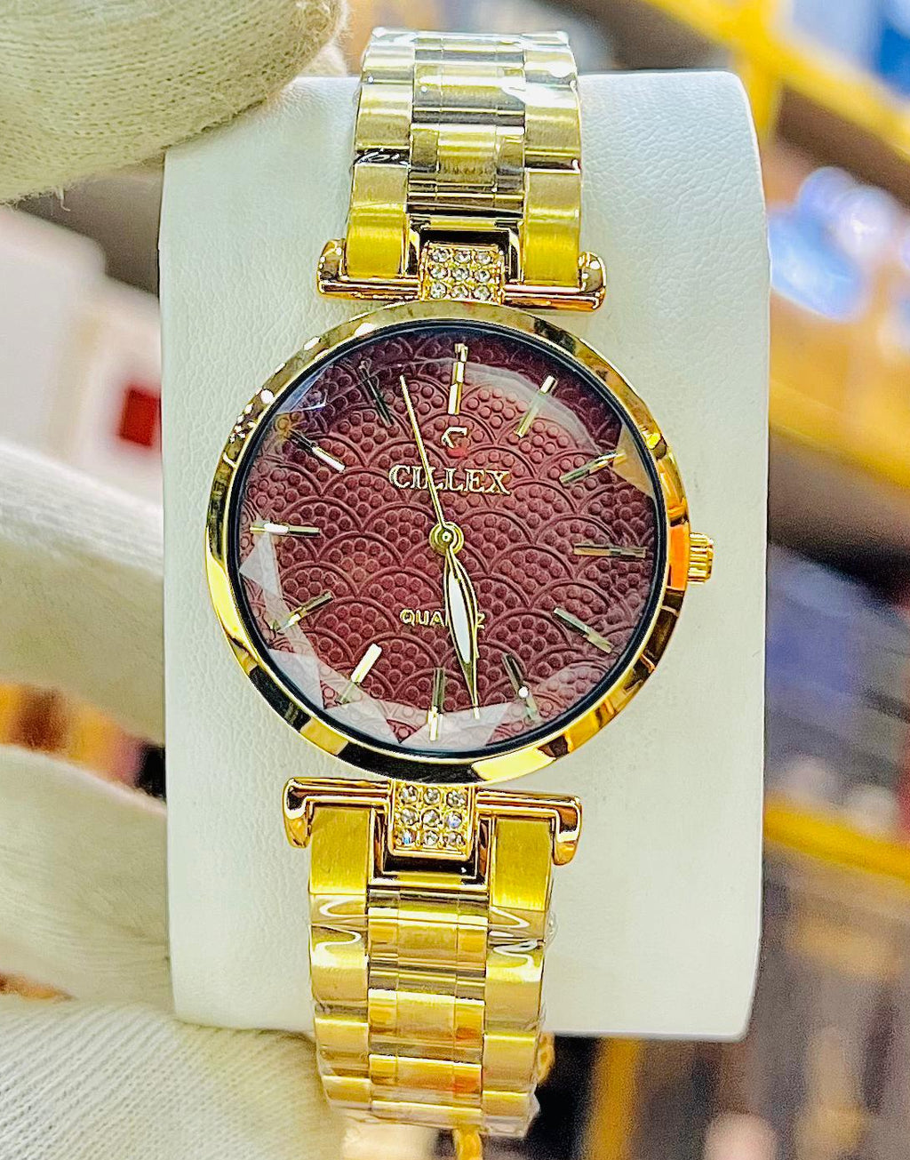 A gold-tone wristwatch with a burgundy dial is held up by a white gloved hand in front of a blurred retail store background.