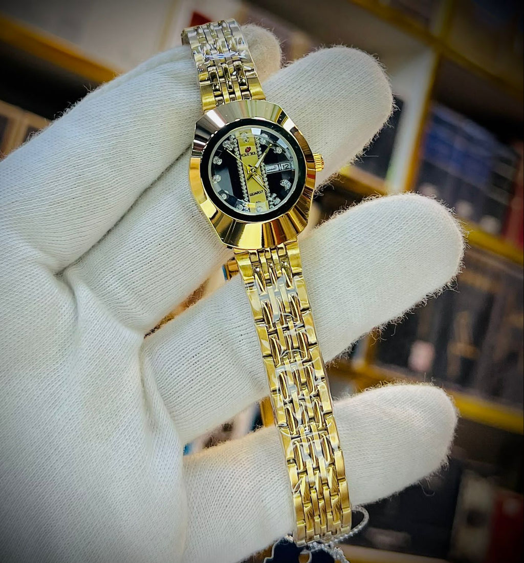 A gold-tone stainless steel watch with a black dial and crystal bezel, held in a white knit-gloved hand. The watch features a two-tone gold and silver link bracelet.
