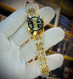 A gold-tone stainless steel watch with a black dial and crystal bezel, held in a white knit-gloved hand. The watch features a two-tone gold and silver link bracelet.